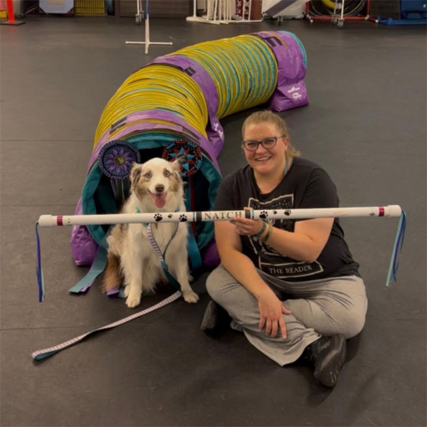 A woman sitting on the floor in front of an agility hurdle with her white dog sitting next to her