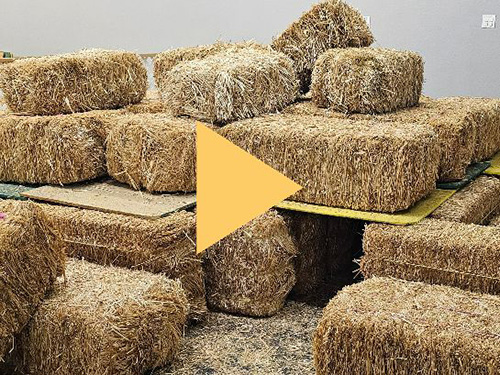 Play button that links to a Facebook video featuring a yellow play arrow overlaying stacks of hay bails.
