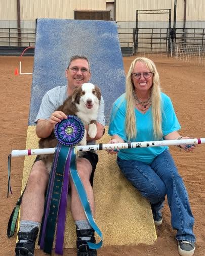 Image of a dog owner and his small brown and white dog posing with a big blue ribbon next to their dog trainer